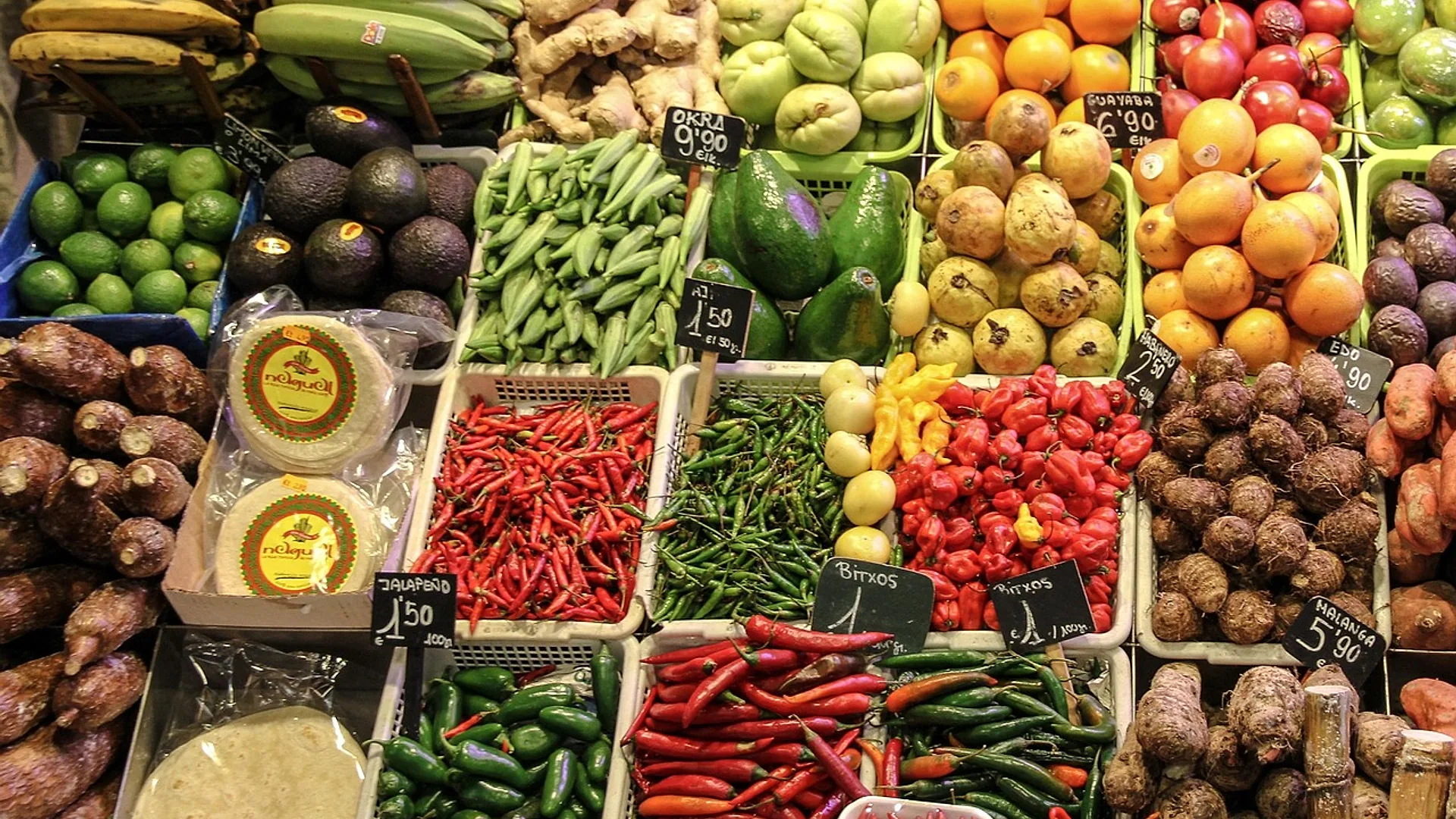 Puesto de mercado visto desde arriba, con cajas de fruta y verdura y pizarras de precios por kilo