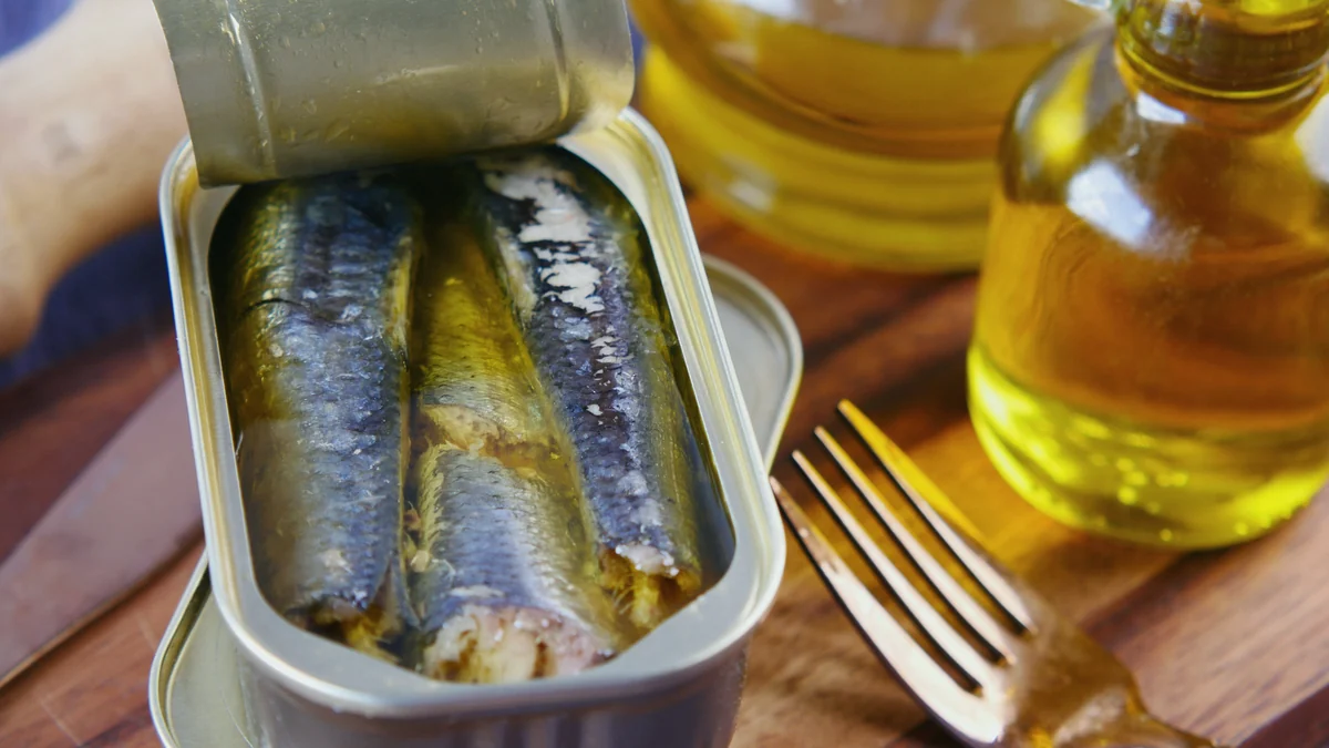 Lata de sardinas en aceite abierta sobre una tabla de madera, con un tenedor y una botella de aceite de oliva