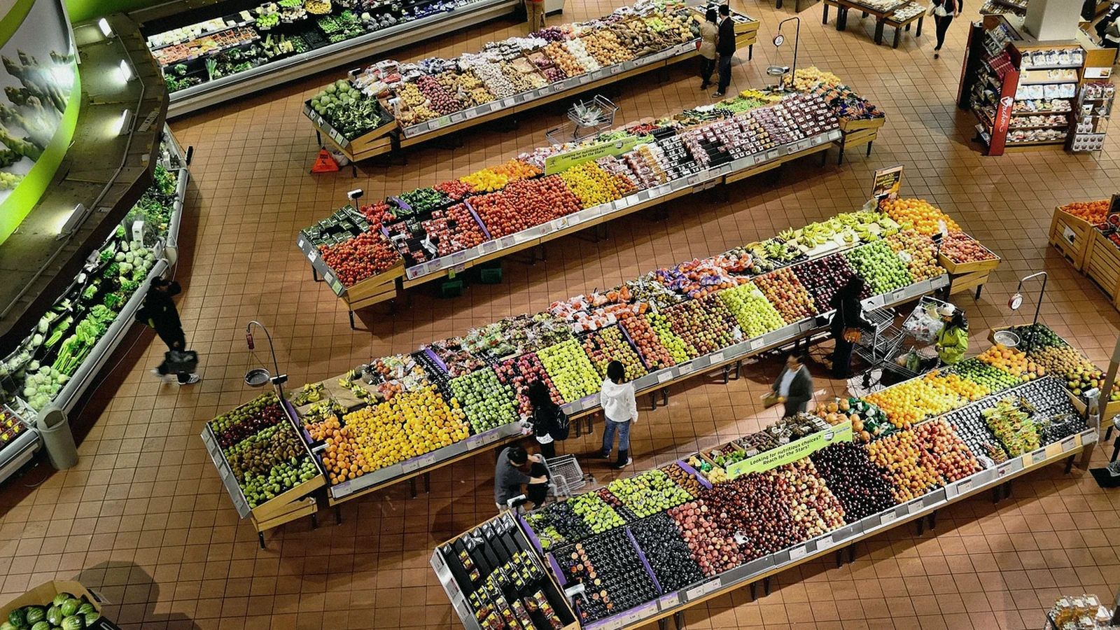 Sección de fruta y verdura de un supermercado vista desde arriba, con varias personas comprando entre los expositores